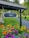 A sign with the text 'Luleå' hangs under a tiled roof structure. Below the sign is a vibrant flower bed with various colorful flowers including orange, yellow, and purple blooms. A gravel path leads into the background where a person can be seen walking under the shade of leafy trees.
