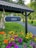 A sign with the text 'Luleå' hangs under a tiled roof structure. Below the sign is a vibrant flower bed with various colorful flowers including orange, yellow, and purple blooms. A gravel path leads into the background where a person can be seen walking under the shade of leafy trees.