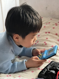A young child wearing a light blue sweater is lying on a polka dot patterned bed. The child is focused on holding and playing with a small, blue handheld electronic game device that has yellow buttons.