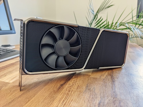 A large, modern graphics card with a bold design sitting on a wooden surface. The card features a prominent cooling fan and a sleek metal frame. In the background, a potted plant adds a touch of greenery and there is a partially visible computer monitor and keyboard.