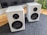 Two white speakers are placed on a grey mat with a wooden surface underneath. The speakers have a sleek and modern design, featuring dual drivers.