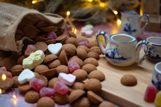 A rustic burlap sack spills a generous amount of small brown cookies onto a wooden surface. Interspersed among the cookies are colorful candies, including gummy shapes and marshmallow hearts. To the right, a set of blue and white ceramic teapots and cups is positioned on a wooden board. Soft golden holiday lights twinkle in the background, creating a warm and cozy atmosphere.