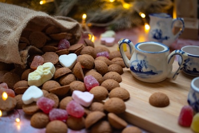 A rustic burlap sack spills a generous amount of small brown cookies onto a wooden surface. Interspersed among the cookies are colorful candies, including gummy shapes and marshmallow hearts. To the right, a set of blue and white ceramic teapots and cups is positioned on a wooden board. Soft golden holiday lights twinkle in the background, creating a warm and cozy atmosphere.