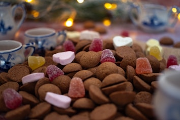 An assortment of small, round, brown cookies topped with colorful gummy candies and marshmallows arranged on a table. In the background, there are blue and white teacups and teapots, alongside some blurred out, warm fairy lights creating a cozy and festive atmosphere.