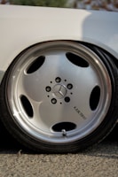 A close-up view of a shiny metallic car wheel featuring the Mercedes-Benz logo in the center. The wheel has a sleek, polished silver finish with five circular openings. The word 'LARZON' is inscribed near the center of the wheel. The reflection on the wheel surface indicates a sunny or brightly lit environment.