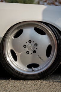 A close-up view of a shiny metallic car wheel featuring the Mercedes-Benz logo in the center. The wheel has a sleek, polished silver finish with five circular openings. The word 'LARZON' is inscribed near the center of the wheel. The reflection on the wheel surface indicates a sunny or brightly lit environment.