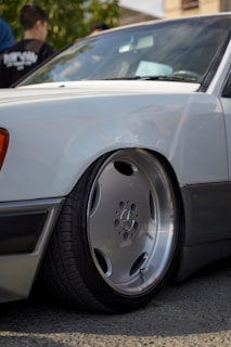A close-up of a white car showcasing its custom lowered suspension and large, aftermarket Mercedes-Benz wheel with a low-profile tire. In the background, there are some people standing, slightly out of focus.