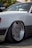 A close-up of a white car showcasing its custom lowered suspension and large, aftermarket Mercedes-Benz wheel with a low-profile tire. In the background, there are some people standing, slightly out of focus.