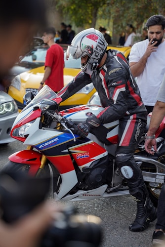 A motorcyclist in full protective gear is seated on a Honda CBR motorcycle, surrounded by onlookers. The motorbike showcases red, white, and blue colors with branding. The background features several people and parked cars, suggesting a gathering or event. One individual appears to be using a smartphone.