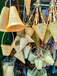 A collection of woven bamboo baskets in various shapes and sizes is displayed, hanging from wires and resting on surfaces. The baskets exhibit intricate weaving patterns, with several conical and cylindrical designs. Wooden and bamboo elements add to the rustic look of the items, and the baskets are densely arranged, overlapping each other.