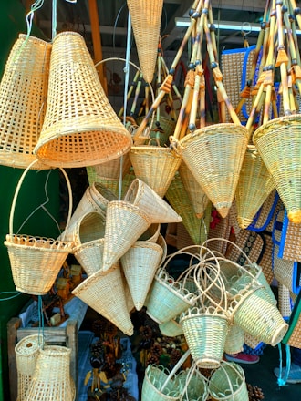A collection of woven bamboo baskets in various shapes and sizes is displayed, hanging from wires and resting on surfaces. The baskets exhibit intricate weaving patterns, with several conical and cylindrical designs. Wooden and bamboo elements add to the rustic look of the items, and the baskets are densely arranged, overlapping each other.