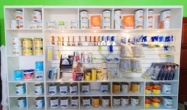 A white shelving unit displays an assortment of paint-related supplies. The top shelves hold various cans of paint with brands like Glasurit. Below are sections filled with paintbrushes, rollers, masking tape, and other painting tools, neatly organized.