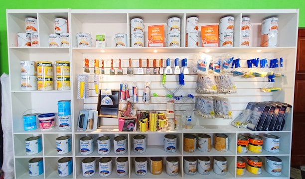 A white shelving unit displays an assortment of paint-related supplies. The top shelves hold various cans of paint with brands like Glasurit. Below are sections filled with paintbrushes, rollers, masking tape, and other painting tools, neatly organized.