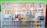 A white shelving unit displays an assortment of paint-related supplies. The top shelves hold various cans of paint with brands like Glasurit. Below are sections filled with paintbrushes, rollers, masking tape, and other painting tools, neatly organized.