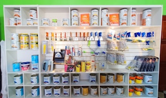 A white shelving unit displays an assortment of paint-related supplies. The top shelves hold various cans of paint with brands like Glasurit. Below are sections filled with paintbrushes, rollers, masking tape, and other painting tools, neatly organized.