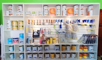 A white shelving unit displays an assortment of paint-related supplies. The top shelves hold various cans of paint with brands like Glasurit. Below are sections filled with paintbrushes, rollers, masking tape, and other painting tools, neatly organized.