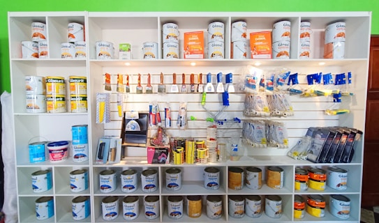 A white shelving unit displays an assortment of paint-related supplies. The top shelves hold various cans of paint with brands like Glasurit. Below are sections filled with paintbrushes, rollers, masking tape, and other painting tools, neatly organized.