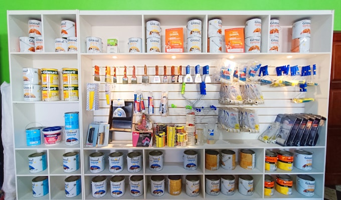 A white shelving unit displays an assortment of paint-related supplies. The top shelves hold various cans of paint with brands like Glasurit. Below are sections filled with paintbrushes, rollers, masking tape, and other painting tools, neatly organized.