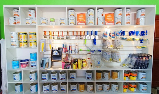 A white shelving unit displays an assortment of paint-related supplies. The top shelves hold various cans of paint with brands like Glasurit. Below are sections filled with paintbrushes, rollers, masking tape, and other painting tools, neatly organized.