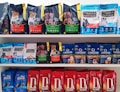 A variety of dog food bags arranged on shelves. The top shelf displays brands such as Cibau and Formula Natural with packaging in black, green, and blue colors. The middle shelf includes Formula Natural and Origens with a mix of blue and red packages. The bottom shelf features Daldog with red packaging.
