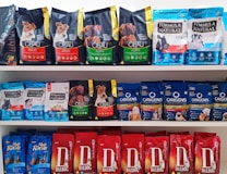 A variety of dog food bags arranged on shelves. The top shelf displays brands such as Cibau and Formula Natural with packaging in black, green, and blue colors. The middle shelf includes Formula Natural and Origens with a mix of blue and red packages. The bottom shelf features Daldog with red packaging.