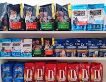 A variety of dog food bags arranged on shelves. The top shelf displays brands such as Cibau and Formula Natural with packaging in black, green, and blue colors. The middle shelf includes Formula Natural and Origens with a mix of blue and red packages. The bottom shelf features Daldog with red packaging.