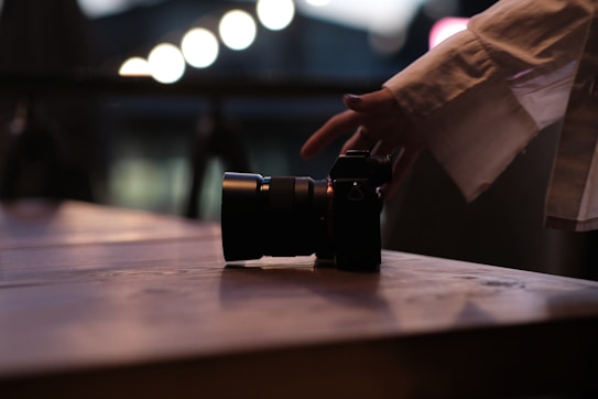A hand reaching towards a camera resting on a wooden table. The scene captures a moment of interaction, set against a soft bokeh background with blurred lights.
