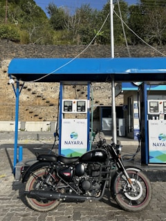 A black Royal Enfield motorcycle is parked in front of a fuel station with two pumps labeled for diesel and petrol. The station has a blue canopy and is branded with the name 'Nayara'. The background features a stone wall and lush green vegetation.