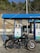 A black Royal Enfield motorcycle is parked in front of a fuel station with two pumps labeled for diesel and petrol. The station has a blue canopy and is branded with the name 'Nayara'. The background features a stone wall and lush green vegetation.