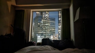 A nighttime cityscape is visible through a window from a dimly lit interior. The foreground features a bed with white bedding and several pieces of luggage next to it. Outside, several tall, illuminated office buildings with many lit windows can be seen against the dark night sky.