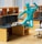 An office workspace with multiple cubicles, dual monitors on the desks, and a white ergonomic office chair. On one of the desks, there are alphabet blocks spelling the name 'FRED'. Above the desk, a blue statue of a human figure is depicted in a dynamic, jumping pose. The workspace shelves contain various office supplies, including stacked books and a clock.