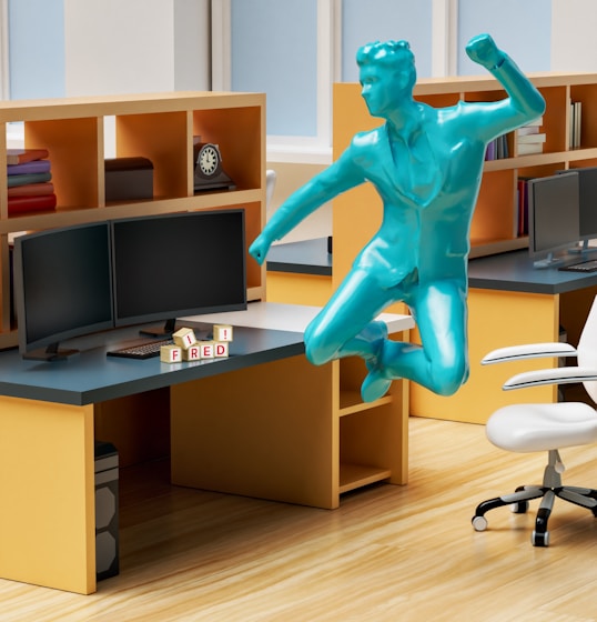 An office workspace with multiple cubicles, dual monitors on the desks, and a white ergonomic office chair. On one of the desks, there are alphabet blocks spelling the name 'FRED'. Above the desk, a blue statue of a human figure is depicted in a dynamic, jumping pose. The workspace shelves contain various office supplies, including stacked books and a clock.