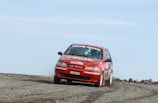 A red rally car is racing on a gravel track, kicking up dust behind it. The car features several sponsor logos and decals, including 'OZUCAB RACING' on the windshield. The environment appears remote with clear skies.