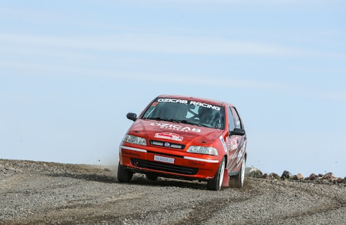 A red rally car is racing on a gravel track, kicking up dust behind it. The car features several sponsor logos and decals, including 'OZUCAB RACING' on the windshield. The environment appears remote with clear skies.