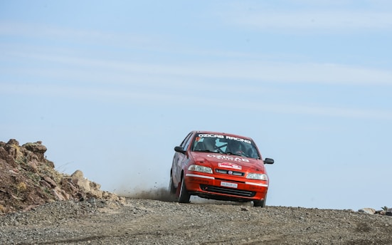 A red rally car is speeding along a rugged, dirt trail. Dust and debris are being kicked up behind it. The vehicle is prominently displaying sponsor logos and racing decals. The setting is an open, barren landscape with clear skies.