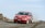 A red rally car with 'Ozicab Racing' written on the windshield drives on a gravel road, kicking up dust behind it. The car is adorned with various decals and has white wheels. The background features a clear sky and rocky terrain.