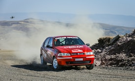 A red rally car speeds on a dirt road, kicking up a trail of dust behind it as it navigates a sharp turn. The rugged terrain is set against a backdrop of hills and open sky. A drone is visible in the sky capturing the action.