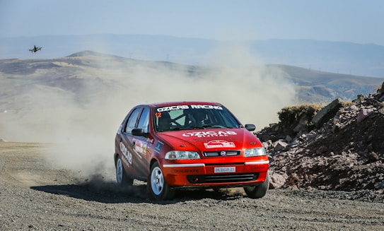 A red rally car speeds on a dirt road, kicking up a trail of dust behind it as it navigates a sharp turn. The rugged terrain is set against a backdrop of hills and open sky. A drone is visible in the sky capturing the action.