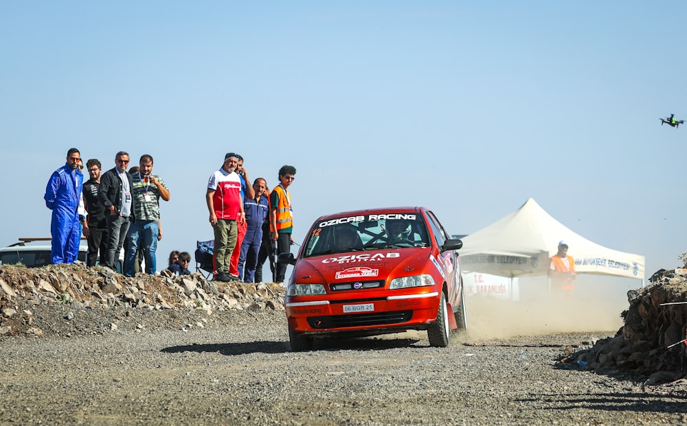 a red car driving down a dirt road next to a group of people