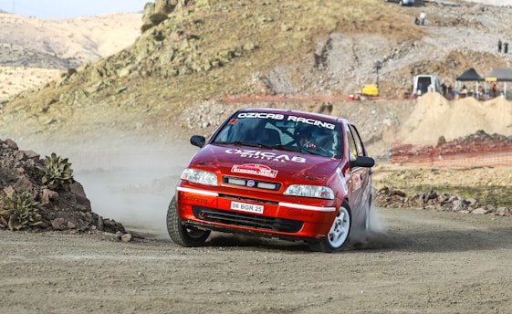 A red rally car is speeding along a dirt track, kicking up dust as it navigates a sharp turn. The vehicle is adorned with various racing decals and has white rims, indicating its involvement in a motor race. The background shows a rugged, rocky landscape with a few small plants and a hill. Some temporary structures and vehicles are visible further along the path, suggesting a racing event setup.