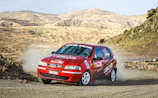 Side view of a rugged rally car kicking up red dirt on a dusty Cambodian trail.