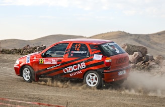 A rally car with red and black stripes is speeding on a dirt road, surrounded by a rocky landscape. Dust trails behind the car as it navigates the terrain. The car has various decals and sponsor logos visible on its body.