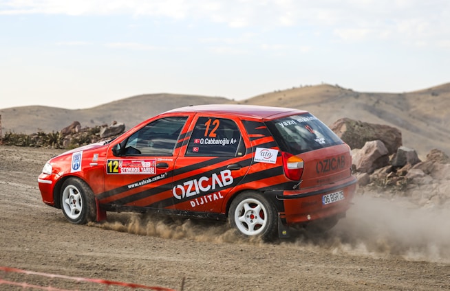 A rally car with red and black stripes is speeding on a dirt road, surrounded by a rocky landscape. Dust trails behind the car as it navigates the terrain. The car has various decals and sponsor logos visible on its body.