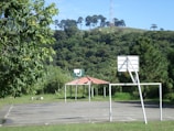 A basketball court is situated outdoors, surrounded by lush green trees and a hillside covered with dense foliage. A basketball hoop without a net stands in the foreground, with a white metal frame and a backboard. In the background, a small structure with a red roof is visible amidst the greenery.
