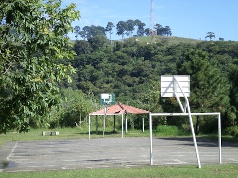 A basketball court is situated outdoors, surrounded by lush green trees and a hillside covered with dense foliage. A basketball hoop without a net stands in the foreground, with a white metal frame and a backboard. In the background, a small structure with a red roof is visible amidst the greenery.