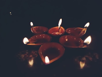 A group of six small clay oil lamps are arranged on a flat surface, each with a burning flame. The dimly lit background accentuates the warm glow from the lamps, casting a soft and serene ambiance.