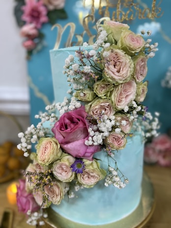 A beautifully decorated two-tier cake with a light blue frosting is adorned with a cascade of roses and other flowers in shades of pink, pale green, and white. Delicate baby's breath flowers are interspersed among the roses, enhancing the elegant appearance. In the background, there are additional floral decorations and golden script on a blue backdrop.