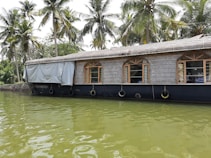 A traditional houseboat with a thatched roof floats on a calm body of water, surrounded by lush green palm trees. The reflection of the boat is visible in the water, creating a serene and picturesque scene. The windows of the boat have wooden frames, adding to its rustic charm.