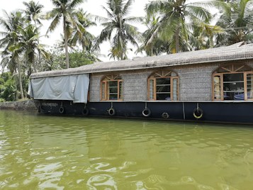 A traditional houseboat with a thatched roof floats on a calm body of water, surrounded by lush green palm trees. The reflection of the boat is visible in the water, creating a serene and picturesque scene. The windows of the boat have wooden frames, adding to its rustic charm.