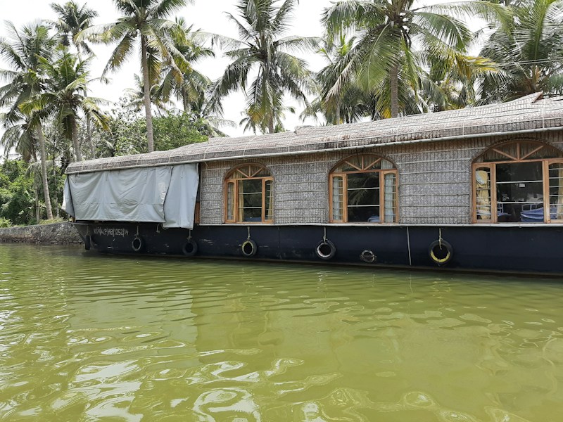 A traditional houseboat with a thatched roof floats on a calm body of water, surrounded by lush green palm trees. The reflection of the boat is visible in the water, creating a serene and picturesque scene. The windows of the boat have wooden frames, adding to its rustic charm.